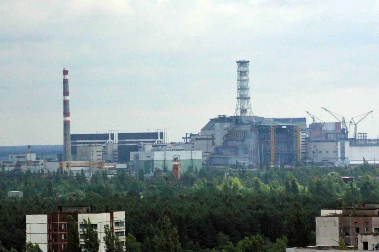 Widok na elektrownię jądrową, która przypomina, gdzie leży Czarnobyl. Widać kominy i budynki przemysłowe otoczone lasem.