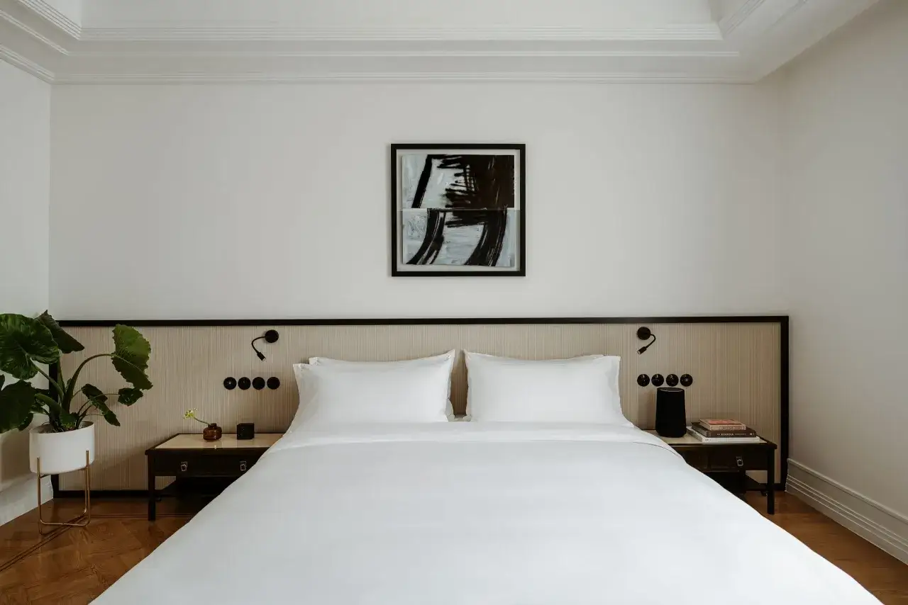 Elegancka sypialnia w hotelu Nobu Warszawa z białym pościelonym łóżkiem, nowoczesnym obrazem i rośliną doniczkową.