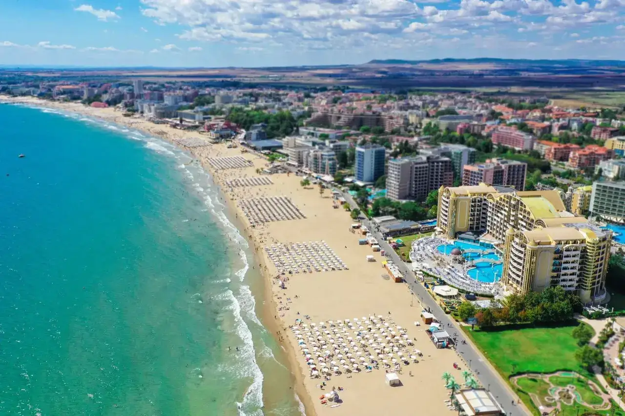 Słoneczny brzeg w Bułgarii, z turkusowym morzem, piaszczystą plażą pełną leżaków i parasoli, oraz luksusowym hotelem z basenami.