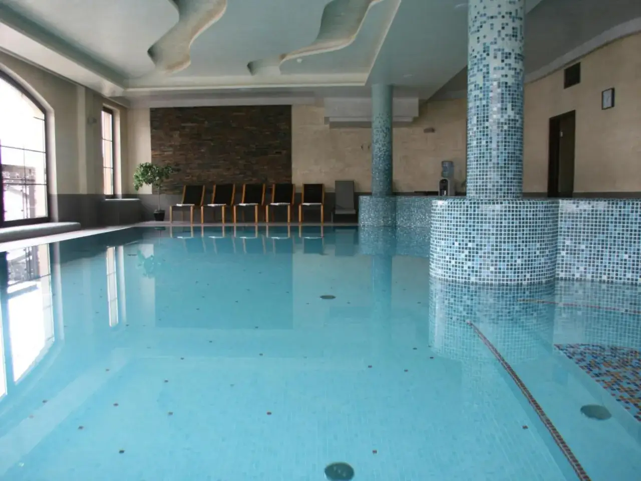 Basen w Hotelu Elbrus Spa & Wellness. Czysta woda, płytki, leżaki i kolumna w mozaice.