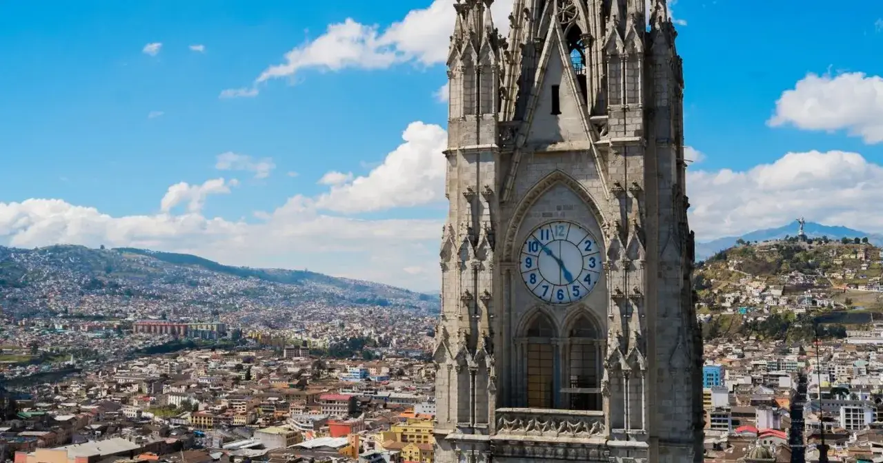 Wieża z zegarem góruje nad Quito, stolicą Ekwadoru. Miasto rozciąga się pośród wzgórz, a w oddali widać posąg na szczycie.