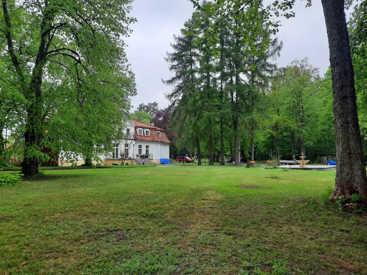 Biały dworek z czerwonym dachem otoczony zielenią. Na trawniku fontanna, drzewa i ławka. Miejsce jak z bajki, idealne na odpoczynek w gierłoż.