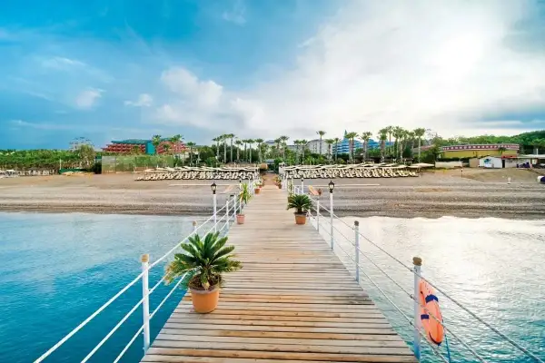 Drewniane molo prowadzi do plaży z rzędami leżaków i palm. W tle widać hotel, który zachęca do sprawdzenia mukarnas spa resort opinie.