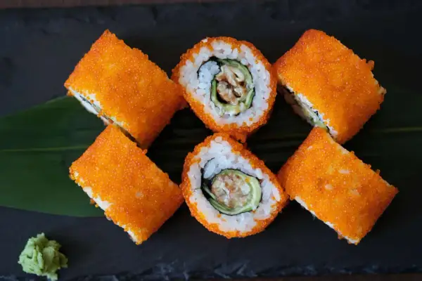 Roladki sushi California z pomarańczowym tobiko, ryżem, awokado i krewetką. Obok zielony listek i odrobina wasabi.