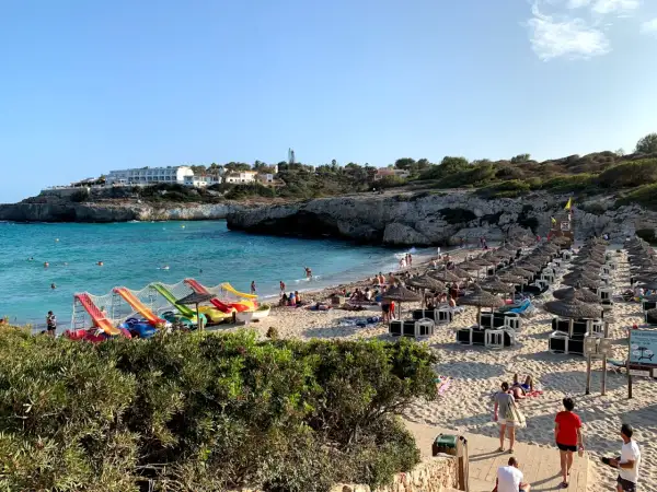 Słoneczna plaża Cala Domingos tętni życiem. Woda kusi turkusowym blaskiem, a piasek ugina się pod ciężarem leżaków i parasoli. Dzieci bawią się na kolorowych zjeżdżalniach.