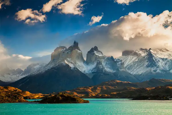 Majestatyczne szczyty Torres del Paine w **Tierra del Fuego** oświetlone słońcem, z turkusowym jeziorem u podnóża.