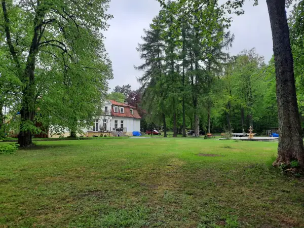 Biały dworek z czerwonym dachem otoczony zielenią. W oddali widać fontannę i drzewa. To urokliwa **gierłoż**.