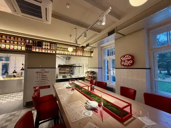 Wnętrze restauracji z czerwonymi krzesłami i długim stołem. Na ścianie wisi neon "Tutto Posto", a nad ladą półki z produktami. Otto Pompieri opinie: warto odwiedzić!