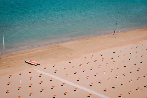 Pusta plaża w Rimini z rzędem pomarańczowych parasoli i małą łodzią. Woda jest turkusowa.