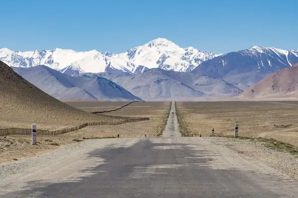 Długa, prosta droga prowadzi przez pustynny krajobraz w kierunku ośnieżonych gór. To widok na Pamir Highway, zapierający dech w piersiach szlak podróżniczy.