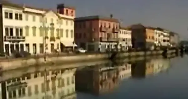 Nadbrzeże z kolorowymi kamienicami odbija się w spokojnej wodzie. W oddali widać fragmenty budynków nad rzeką.