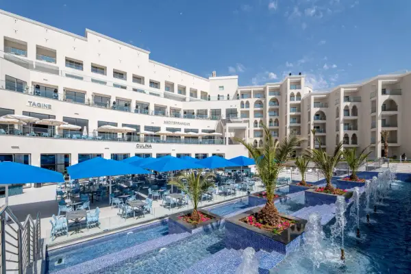 Luksusowy hotel z basenem i restauracją na świeżym powietrzu. Widać palmy, niebieskie parasole i stoły z krzesłami. Idealne miejsce na wakacje, jak na zdjęciach z **pickalbatros**.